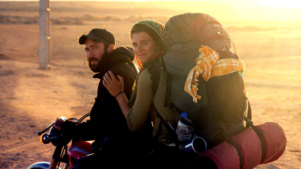Eine Frau mit Rucksack und ein Mann mit Bart sitzen auf einem Motorrad, das vor einem weiten Feld bei Sonnenuntergang steht. 