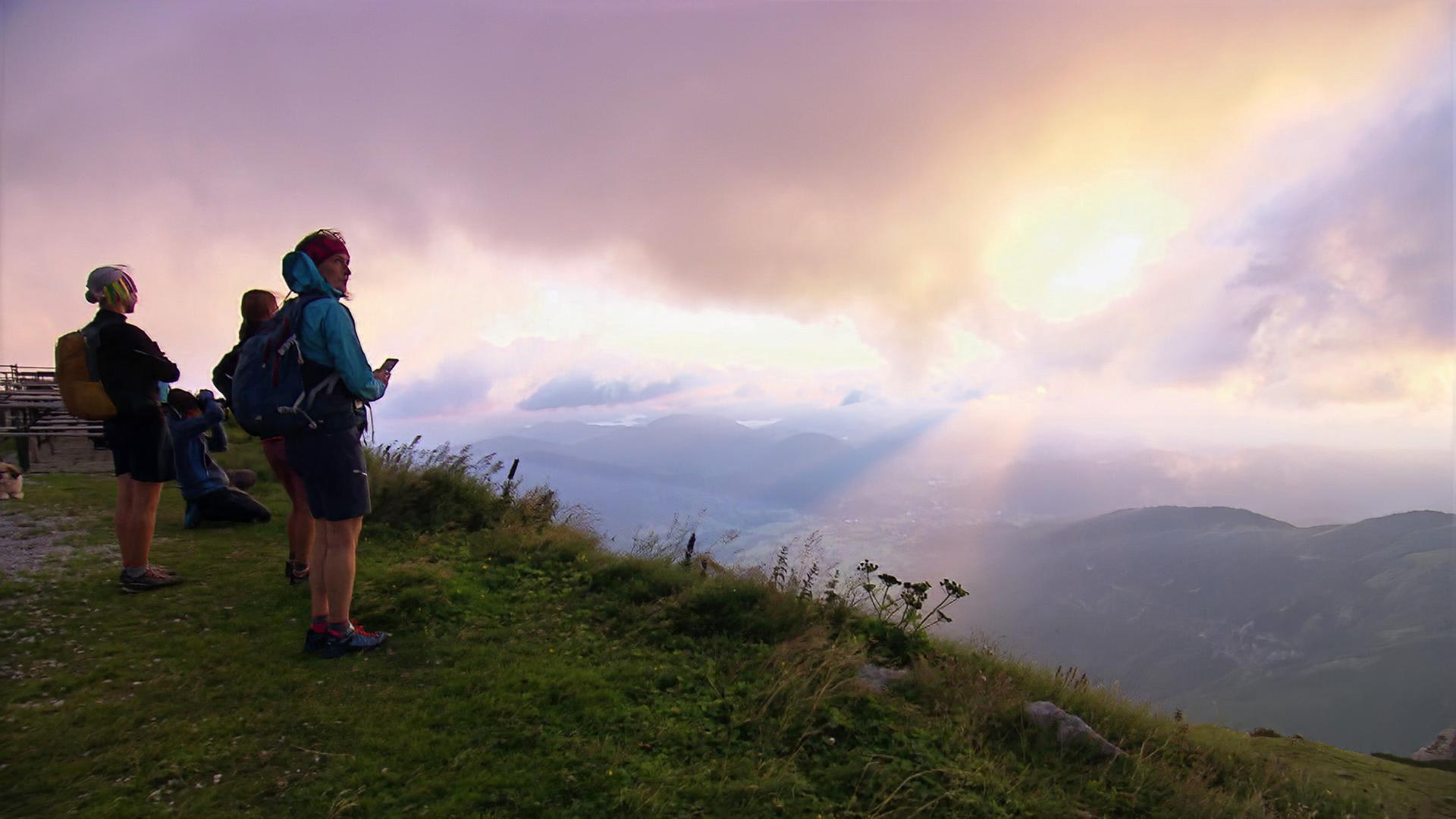 Die Darstellung zeigt eine Gruppe von Wanderern an einem Berg, wahrscheinlich in Niederösterreich. Im Vordergrund steht eine Person in Wanderkleidung mit Rucksack, die in die Ferne schaut und möglicherweise ein Smartphone in der Hand hält. Weitere Wanderer stehen daneben und beobachten die Aussicht. Der Hintergrund ist von dramatischen Wolken geprägt, durch die Lichtstrahlen scheinen. Die Landschaft ist hügelig und grün, mit einer Atmosphäre von Weite und Erhabenheit.   Die Aufnahme scheint während der goldenen Stunde gemacht worden zu sein, da eine warme Lichtstimmung herrscht. Die Szenerie vermittelt ein Gefühl der Erkundung und des Abenteuers in der Natur.