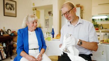Eine ältere, sitzende Frau schaut einem älteren Mann mit Brille und weissen Handschuhen zu, wie er einen silbernen Kerzenleuchter mit einem weissen Tuch abwischt.