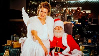 Weihnachtsmann und Christkind sitzen auf einem Sessel, hinter ihnen Weihnachtsdeko und Geschenke