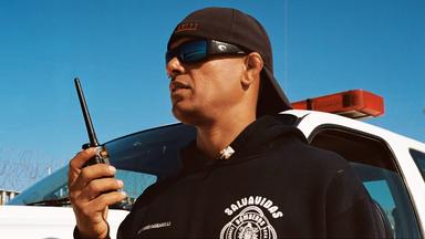 Rettungsschwimmer mit Sonnenbrille, Baseball-Kappe und Funkgerät vor einem Einsatzfahrzeug mit Sirene, im Hintergrund ein Metallzaun mit Stacheldraht.