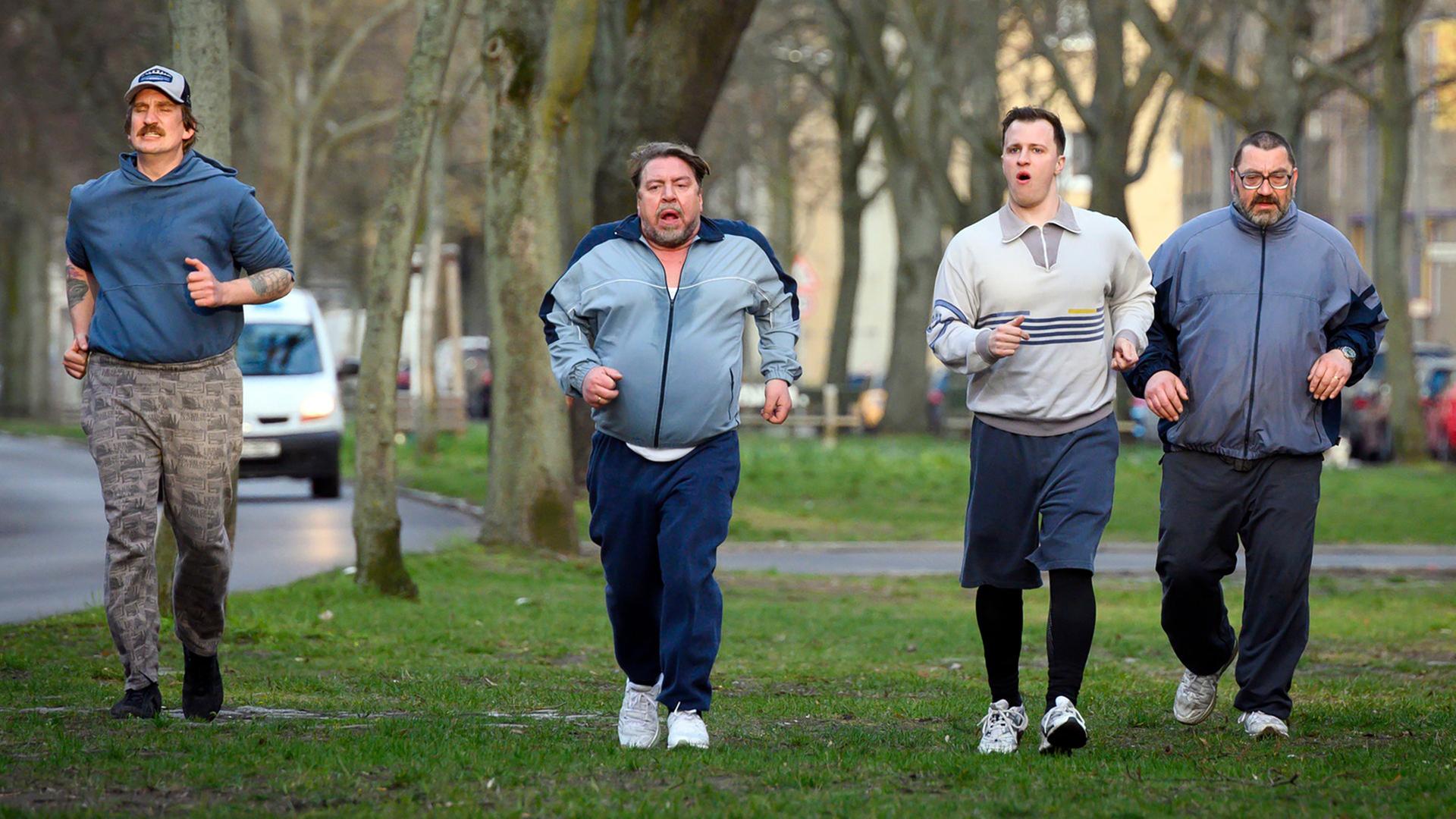 Vier Männer in Sportkleidung joggen angestrengt unter Bäumen auf einer Wiese.