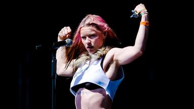 Eine leicht bekleidete Sängerin mit roten Strähnen in den Haaren lässt auf der Bühne mit erhobenen Armen die Oberarmmuskeln spielen.