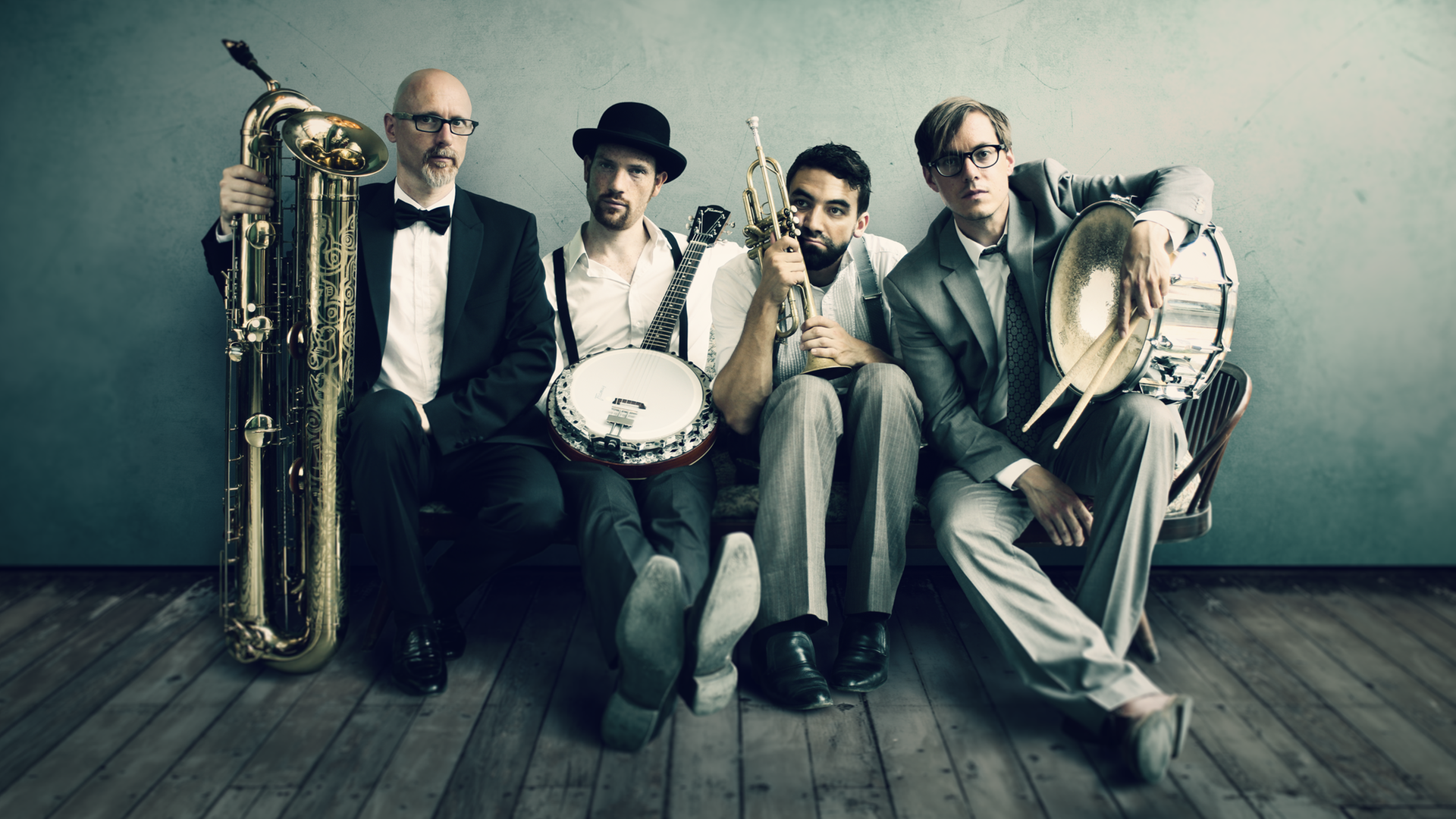 Vier Männer sitzen auf einem Holzboden an einer Wand und halten Musikinstrumente in der Hand.