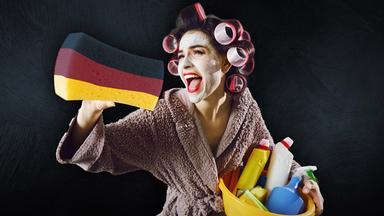 Eine Frau im Bademantel mit Lockenwicklern im Haar, einem Putzeimer mit vielen Putzmitteln im Arm und einem großen schwarz/rot/gold eingefärbten Schwamm.