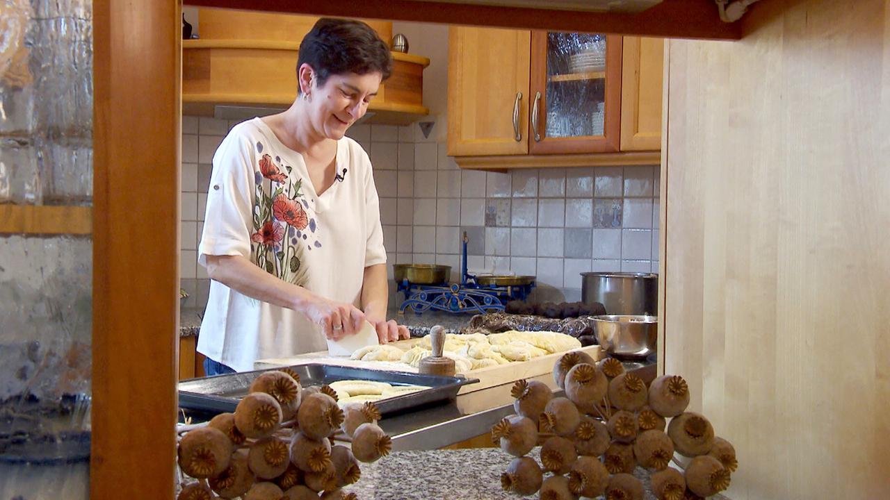 Das Bild zeigt eine Küchenszene, in der eine Frau bei der Zubereitung von Backwaren beschäftigt ist. Sie steht an einer Arbeitsfläche aus grauem Granit und schneidet mit einem Schneider ein Stück Teig. Die Frau trägt ein helles T-Shirt mit floralem Muster und hat kurze, dunkle Haare. Im Hintergrund sind verschiedene Küchenelemente sichtbar, darunter ein Herd mit Töpfen und eine glasierte Fliese an der Wand.  Auf der Arbeitsfläche sind verschiedene Backwaren in Form von Teiglingen zu sehen, die darauf warten, weiterverarbeitet zu werden. Vor ihr steht eine Backform. Im Vordergrund des Bildes sind Mohnkapseln platziert, die zur Tradition des Mohnanbaus in der Region gehören und eine Verbindung zur Zubereitung von Mohngebäck herstellen. Das gesamte Setting vermittelt eine gemütliche, heimelige Atmosphäre, die typisch für das Backen in der Vorweihnachtszeit ist.