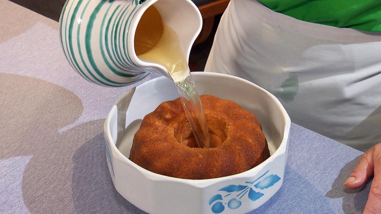 Das Bild zeigt eine Person, die einen hellen, flüssigen Inhalt aus einer Kanne in eine runde Kuchenform gießt. Die Form hat eine konzentrische Vertiefung in der Mitte und ist mit einer goldbraunen Kuchenoberfläche gefüllt. Der Tisch ist mit einer hellblauen Stoffdecke bedeckt. Die Kanne ist weiß mit grünen Streifen und hat eine runde Form. Die Person ist mit einem grünen Oberteil und einer weißen Schürze bekleidet. Das Bild vermittelt die Atmosphäre des Backens und der Zubereitung traditioneller Weihnachtsgerichte.