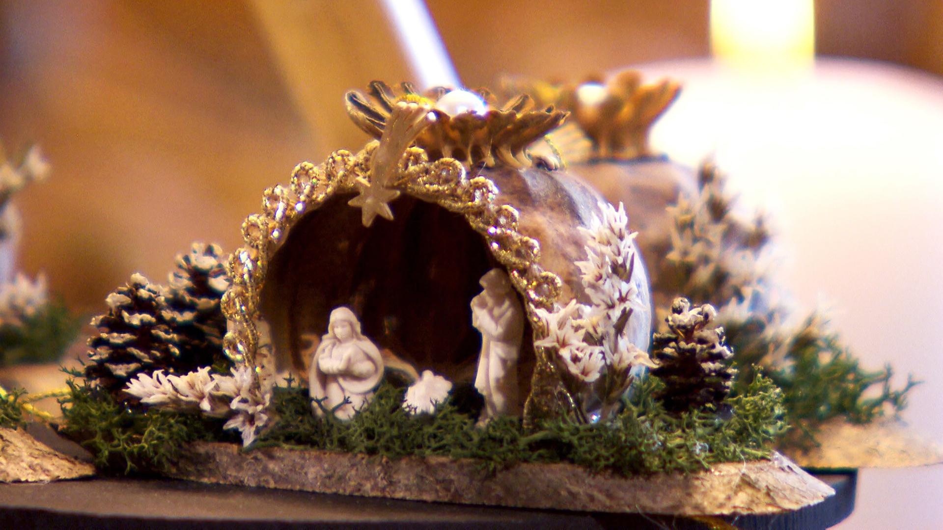 Das Bild zeigt eine kunstvoll gestaltete Krippe, die aus Mohnkapseln gefertigt ist. Die Krippe hat die Form eines kleinen, gewölbten Daches und ist mit goldfarbenen Verzierungen und einem kleinen Stern an der Spitze dekoriert. Vor der Krippe befindet sich eine Darstellung der heiligen Familie, mit Maria, Joseph und dem Jesuskind. Um die Krippe herum sind kleine, kunstvolle Details wie weiße Pflanzen und Zweige sowie Miniatur-Tannenzapfen angeordnet, die die Szenerie lebendig wirken lassen. Im Hintergrund sind weich beleuchtete Kerzen und unscharfe, warme Farben zu erkennen, die eine festliche und ruhige Atmosphäre schaffen. Diese Darstellung vermittelt den besinnlichen Charakter der Advents- und Weihnachtszeit in Österreich.