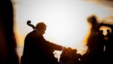 Das Bild zeigt einen Musiker, der ein Cello spielt, während die Sonne am Horizont aufgeht. Die Silhouette des Musikers ist gut erkennbar, er trägt einen dunklen Anzug und hat blondes Haar. Hinter ihm erstreckt sich ein schöner Lichtschein des Sonnenuntergangs, der das Wasser im Hintergrund beleuchtet. Es sind mehrere Menschen in der Nähe des Musikers zu sehen, die in einem gemütlichen Setting zusammensitzen und die Musik genießen. Die Atmosphäre wirkt entspannt und festlich, passend zu einem musikalischen Ereignis im Freien. 