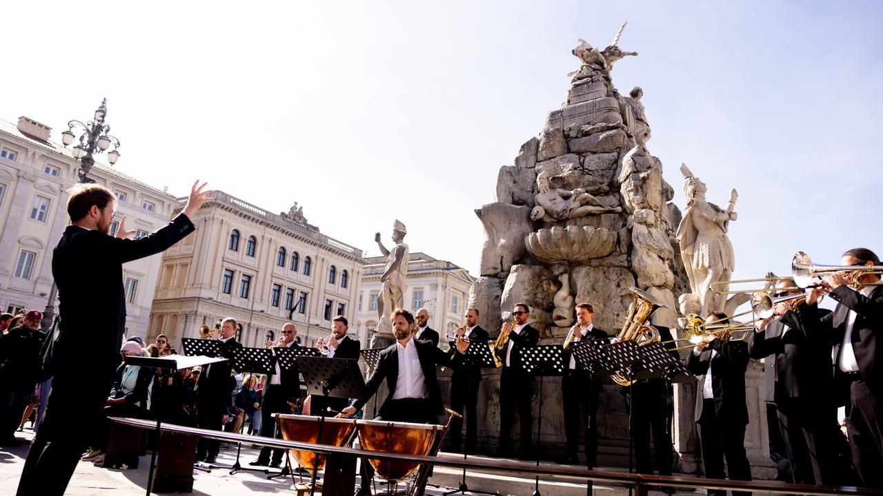 Das Bild zeigt eine musikalische Darbietung im Freien. Im Vordergrund steht ein Dirigent, der mit erhobenen Armen das Orchester leitet. Er trägt einen eleganten schwarzen Anzug und hat einen Bart. Vor ihm stehen mehrere Musiker, die verschiedene Blasinstrumente spielen. Sie sind ebenfalls formell gekleidet und stehen hinter Notenständern.  Im Hintergrund ist eine große, kunstvolle Statue zu sehen, die Teil eines Brunnens ist. Die Skulptur zeigt Figuren, die in einem historischen Stil gestaltet sind, mit einer Darstellung von einem Krieger und anderen mythologischen Elementen. Rund um den Platz sind historische Gebäude mit hellen Fassaden sichtbar, die zum architektonischen Charme des Ortes beitragen. Der Himmel ist klar und blau, was auf einen sonnigen Tag hinweist.  Die gesamte Szene vermittelt eine festliche Atmosphäre, die mit der musikalischen Aufführung verbunden ist. Das Event findet im Rahmen des "Primavera da Vienna" Festivals statt, und es scheint, dass das Publikum, das sich im Hintergrund versammelt hat, die Aufführung aufmerksam verfolgt.