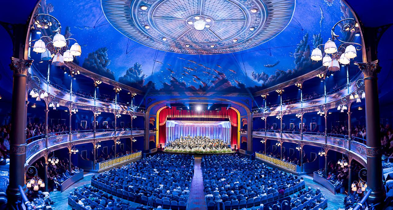 Das Bild zeigt den Innenraum eines Theaters während eines Konzerts. Die Bühne ist festlich dekoriert und wird von einem roten Vorhang eingerahmt, vor dem ein Orchester mit vielen Musikern auftritt. Im Vordergrund sind zahlreiche Reihen eleganter Sitzplätze, die von einer großen Anzahl von Besuchern besetzt sind.

Die Decke des Theaters ist kunstvoll gestaltet und zeigt eine blaue Himmelsszene mit Wolken und Lichtpunkten, die vermutlich Sterne darstellen. Die Wände und die Balkone des Theaters sind reich verziert, und es hängen mehrere elegant gestaltete Lampen, die eine warme, einladende Atmosphäre schaffen.

Es sind auch mehrere Stockwerke mit Sitzplätzen zu sehen, die in einem Halbkreis um die Bühne angeordnet sind. Insgesamt vermittelt das Bild eine festliche Stimmung, passend zu der beschriebenen Veranstaltung, dem traditionellen Osterkonzert der Wiener Symphoniker im Politeama Rossetti in Triest.