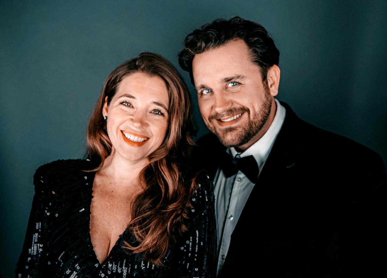 Das Bild zeigt zwei Personen, die dicht nebeneinander stehen und lächeln. Die Frau hat lange, lockige, braune Haare und trägt ein schwarzes, glitzerndes Kleid mit einem tiefen V-Ausschnitt. Sie hat rote Lippenstiftfarbe aufgetragen und ihr Gesicht strahlt Freude aus. Der Mann neben ihr hat kurze, dunkle Haare und einen gepflegten Bart. Er trägt ein formelles Outfit, das aus einem schwarzen Sakko und einer weißen Hemdkragen besteht, sowie eine schwarze Fliege. Der Hintergrund des Bildes ist in einem sanften, einheitlichen Blauton gehalten, der die Personen hervorhebt.