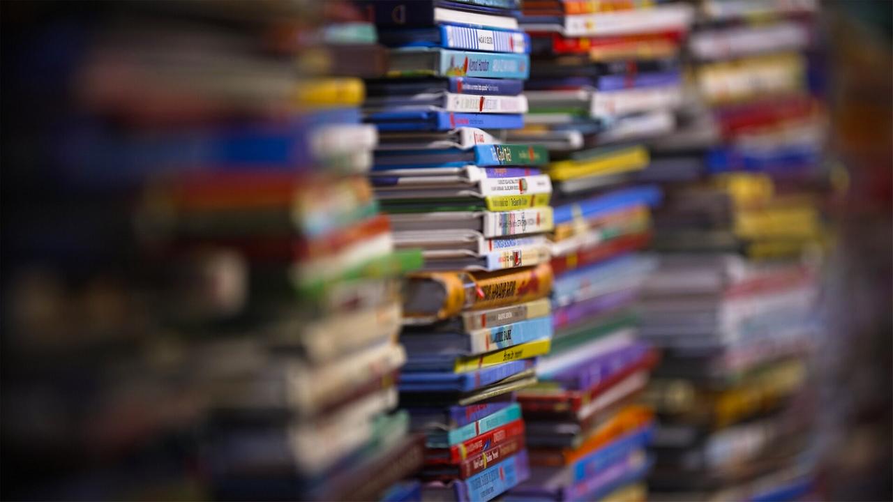 Das Bild zeigt eine Vielzahl von bunten Buchrücken, die übereinander gestapelt sind. Die Bücher sind dicht beieinander angeordnet, wodurch nur die oberen Kanten sichtbar sind. In der Bildmitte sind verschiedene Farben und Größen der Buchrücken erkennbar, überwiegend in Blau, Gelb, Rot und Grün. Der Hintergrund ist unscharf, was die Fokussierung auf die Bücher verstärkt. Diese Darstellung vermittelt den Eindruck einer gut gefüllten Buchhandlung, die eine große Auswahl an Literatur bereithält.