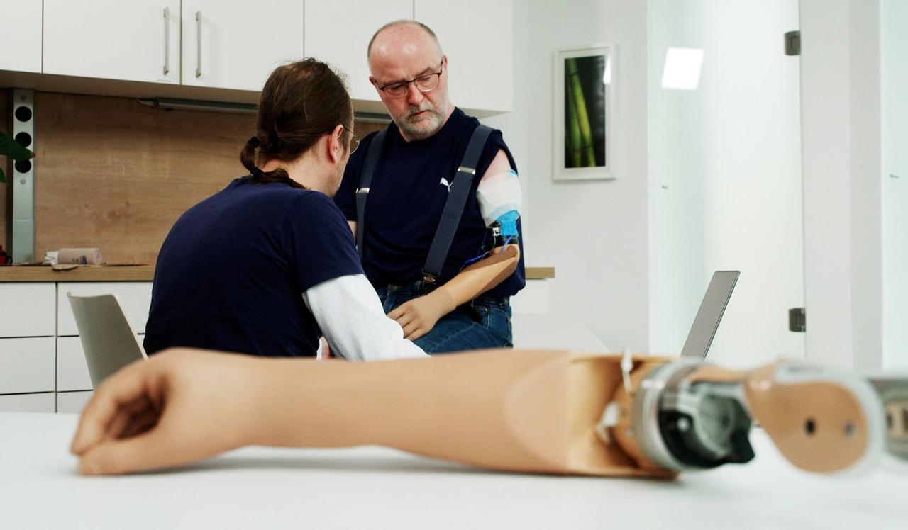 Ein Mann mit Armprothese steht vor einem sitzenden Mann. Im Vordergrund liegt ein Roboterarm.