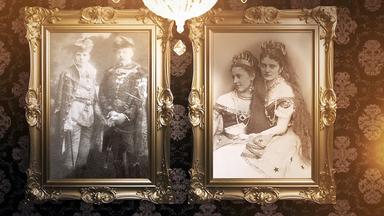 Das Bild zeigt eine ruhige Inneneinrichtung mit zwei gerahmten Fotografien, die an einer Wand hängen. Das linke Foto zeigt einen Mann und eine Frau in formeller Kleidung, wobei der Mann einen Gehstock hält und die Frau eine Tiara trägt. Das rechte Foto zeigt eine Frau in einem langen Kleid und einen Mann im Anzug, beide blicken direkt in die Kamera.