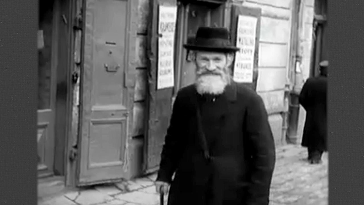 Auf dieser Schwarz-Weiß-Fotografie steht ein bärtiger Mann vor einem Gebäude, gekleidet in einen schwarzen Mantel und Hut. Er hält einen Gehstock in seiner rechten Hand.