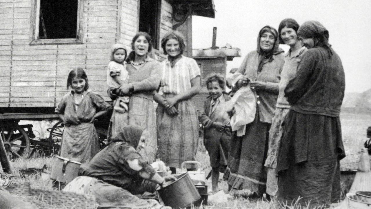 Diese Schwarz-Weiß-Fotografie zeigt eine Gruppe von Frauen und Kindern, die sich vor einer Holzhütte versammelt haben. Eine ältere Frau im Vordergrund rührt in einem Kochtopf.