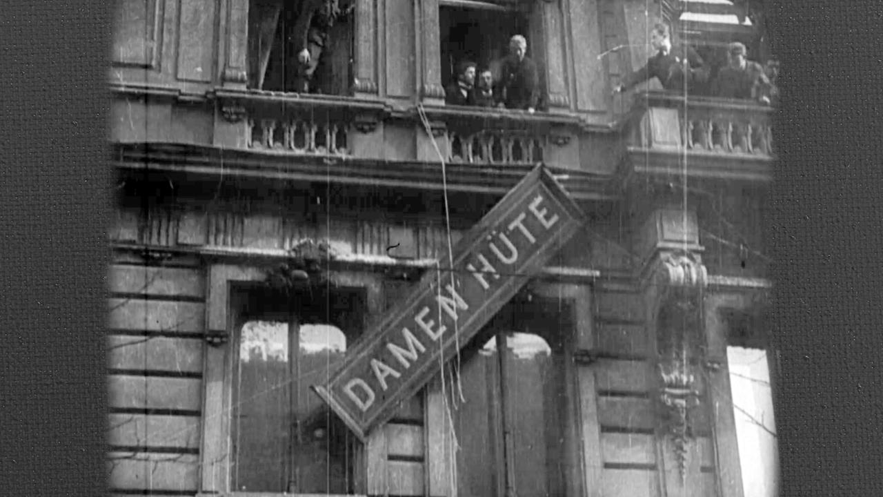 Das Bild zeigt eine Schwarz-Weiß-Szene eines Gebäudes mit einem großen Schild, auf dem "DAMEN HÜTE" steht, das an der Seite des Gebäudes hängt. Das Schild ist an einem Seil herab gelassen. Das Gebäude hat einen Balkon im zweiten Stock, und eine Person steht auf dem Balkon und blickt auf das Schild herab.