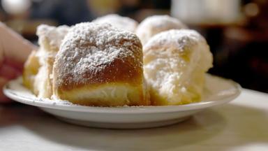 Sechs goldbraune Brötchen auf einem weißen Teller, jedes mit Puderzucker bestäubt, in einem kreisförmigen Muster angeordnet. Der Teller ruht auf einer weißen Tischdecke, und im Hintergrund ist eine verschwommene Hand zu erkennen.