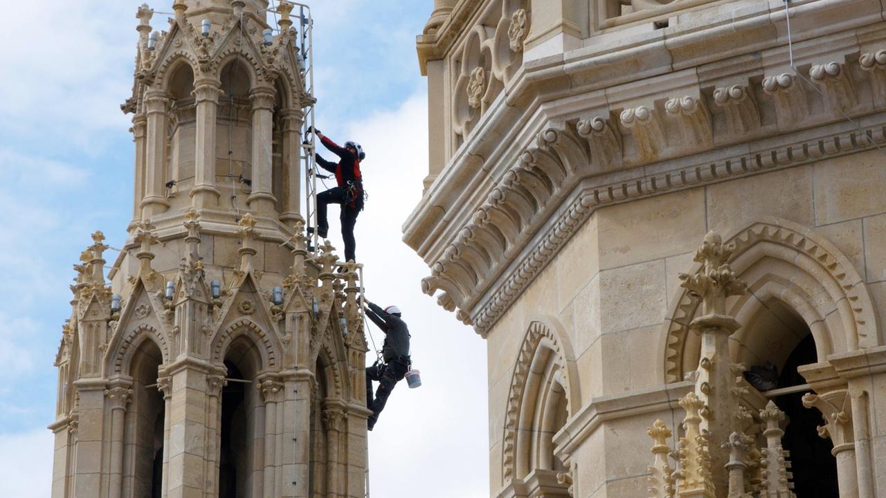 Das Bild zeigt zwei Männer, die an einem hohen Turm arbeiten, der Teil des Wiener Rathauses ist. Der Turm ist reich verziert mit gotischen Elementen und hat verschiedene Architekturdetails, darunter Spitzbögen und Ornamentik. Die Männer sind mit Sicherheitsausrüstung ausgestattet, darunter Helme und Klettergurte, und benutzen eine Leiter, um an die oberen Bereiche des Turms zu gelangen. Im Hintergrund ist ein klarer blauer Himmel mit wenigen Wolken zu sehen. Der Fokus des Bildes liegt auf der Arbeit der Handwerker, die historische Strukturen des Rathauses instandhalten.