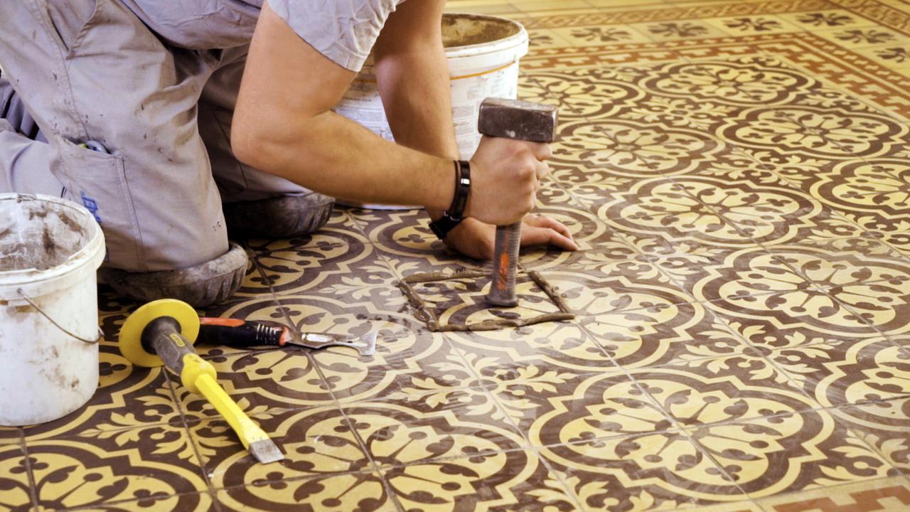 Das Bild zeigt eine Person, die auf dem Boden kniet und eine Art meißelndes Werkzeug in der Hand hält. Sie arbeitet auf einem dekorativen Bodenbelag, der aus quadratischen Fliesen besteht, die mit einem filigranen, floralen Muster in Braun- und Gelbtönen gestaltet sind. Um die Person herum befinden sich Werkzeuge wie ein Hammer, ein Schraubenzieher und eine gelbe Basis (vermutlich ein Werkzeug). Im Hintergrund ist ein Eimer sichtbar, der mit einer unbestimmten Substanz gefüllt ist. Die Szene vermittelt den Eindruck von handwerklicher Arbeit, die Teil der Erhaltung oder Restaurierung des historischen Wiener Rathauses ist.