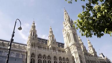 Das Bild zeigt das Wiener Rathaus, ein beeindruckendes neugotisches Gebäude, das sich durch seine aufwändige Architektur auszeichnet. Die Fassade ist aus hellem Stein und wird durch zahlreiche filigrane Verzierungen akzentuiert. Auf beiden Seiten des Rathauses ragen hohe Türme empor, die mit goldenen Zifferblättern und anderen dekorativen Elementen geschmückt sind.   Im Vordergrund ist ein Straßenlaterne zu sehen, die zur städtischen Umgebung gehört. Am oberen Bildrand ist ein klarer Himmel zu erkennen, der das Rathaus in ein freundliches Licht taucht. Im Bild sind auch grüne Äste von Bäumen sichtbar, die dem Motiv eine frische Note verleihen.   Das Rathaus steht zentral in Wien und ist bekannt für seine vielfältigen Veranstaltungen und politischen Funktionen. 