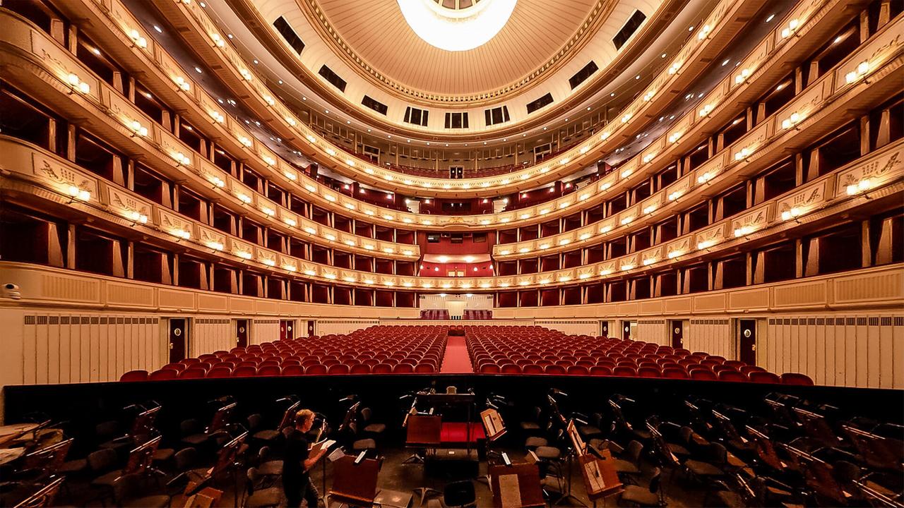 Das Bild zeigt den Innenraum der Wiener Staatsoper aus der Perspektive der Bühne. Im Vordergrund sind Sitzplätze für das Orchester zu sehen, die in einer Reihe angeordnet sind und teilweise mit Notenständern ausgestattet sind.   Hinter den Orchestermusikern erstreckt sich ein großer Zuschauerraum mit roten Samtsitzen, die in geschwungenen Reihen angeordnet sind. Es gibt mehrere Ranglogen, die sich über den gesamten Saal verteilen. Die Wände sind in einem eleganten, hellen Farbton gestaltet und sind mit goldenen Lampen beleuchtet, die eine warme Atmosphäre schaffen.   Direkt über dem Zuschauerraum befindet sich eine große, runde Deckenleuchte, die das gesamte Bild beleuchtet. Die Architektur des Raumes ist geschwungen und vermittelt ein Gefühl von Größe und Pracht. Der gesamte Innenraum strahlt einen historischen Charme aus.   Das Bild ist in warmen Farben gehalten, was die elegante und festliche Stimmung des Ortes unterstreicht.