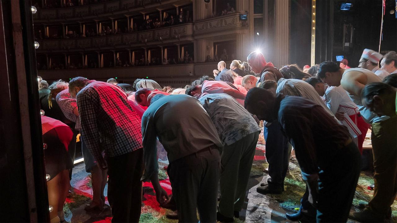Das Bild zeigt eine Szene im Inneren der Wiener Staatsoper. Im Vordergrund sind mehrere Menschen zu sehen, die sich verbeugen. Die Personen tragen unterschiedliche Kleidung; einige haben Karomuster, andere Streifen oder einfarbige Oberteile. Die Haltung der Menschen deutet auf einen Abschlussapplaus hin, wahrscheinlich nach einer Aufführung.   Im Hintergrund sind die Stufen und Sitzreihen des Zuschauerraums sichtbar, die von der Bühne aus betrachtet werden. Das Licht stammt von Scheinwerfern, die eine warme Atmosphäre schaffen. Einige Lichter sind entlang der Seiten sichtbar. Der Boden der Bühne ist unregelmäßig und scheint mit Resten von Bühnenmaterial bedeckt zu sein.  Die Gesamtstimmung des Bildes ist feierlich, und die Umgebung signalisiert, dass die Szene Teil eines kulturellen Ereignisses ist.