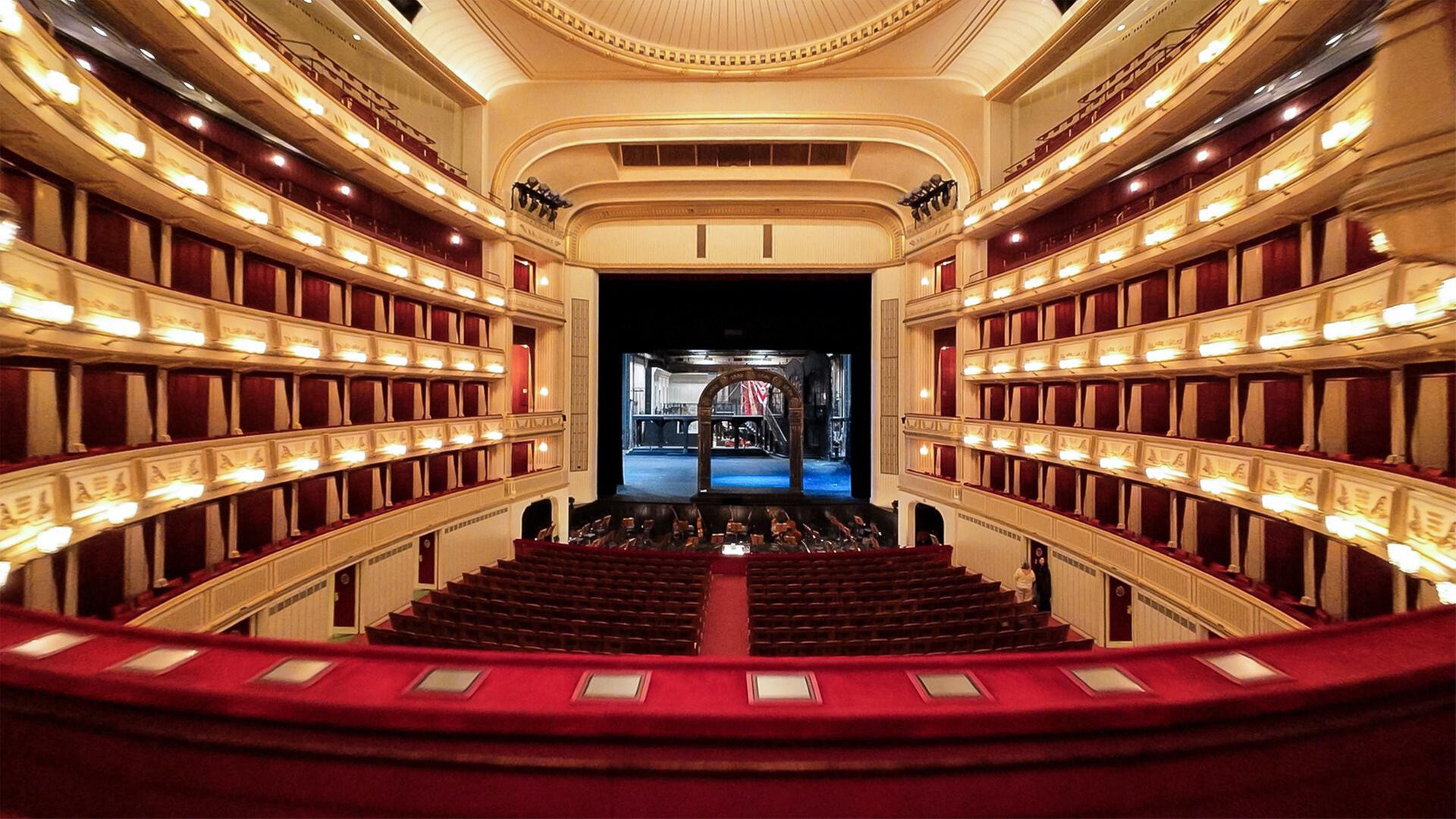 Das Bild zeigt den Innenraum der Wiener Staatsoper. Im Vordergrund sind rote gepolsterte Sitzplätze sichtbar, die in einem leicht geschwungenen Muster angeordnet sind. Über den Sitzplätzen befinden sich Balkone, die mit goldenen Verzierungen und rot-beige gestreiften Vorhängen dekoriert sind. An den Wänden sind mehrere Wandleuchten angebracht, die ein warmes Licht abgeben.  In der Mitte des Bildes ist die Bühne zu sehen, die dunkel und leer ist. Im Hintergrund des Bühnenszenarios sind technische Elemente und ein Teil des Bühnenbildes sichtbar, das eine architektonische Struktur zeigt.   Die gesamte Atmosphäre des Raumes vermittelt ein Gefühl von Eleganz und Tradition. Darüber hinaus kann man die Bühne in ihrer Vorbereitungsphase erkennen, da einige Techniker im Hintergrund zu sehen sind. Der Raum wirkt groß und offen, gekennzeichnet durch hohe Decken und aufwendig gestaltete Elemente.