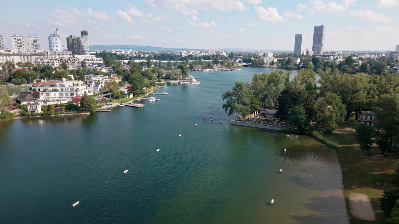 Die Ansicht zeigt die Alte Donau in Wien mit grünen Ufern, Bootsanlegern und Wohnhäusern. Im Hintergrund sind moderne Gebäude sichtbar. Der Himmel ist teilweise bewölkt.