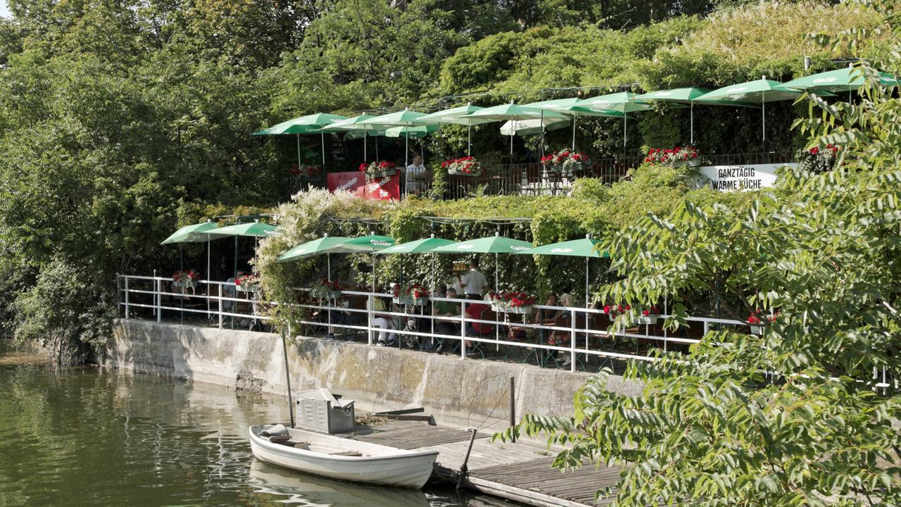 Am Ufer der Alten Donau steht ein Gasthaus mit grünen Sonnenschirmen und blühenden Pflanzen. Ein Boot liegt am Steg, während Gäste im Freien sitzen und essen. Bäume umgeben die Szene.