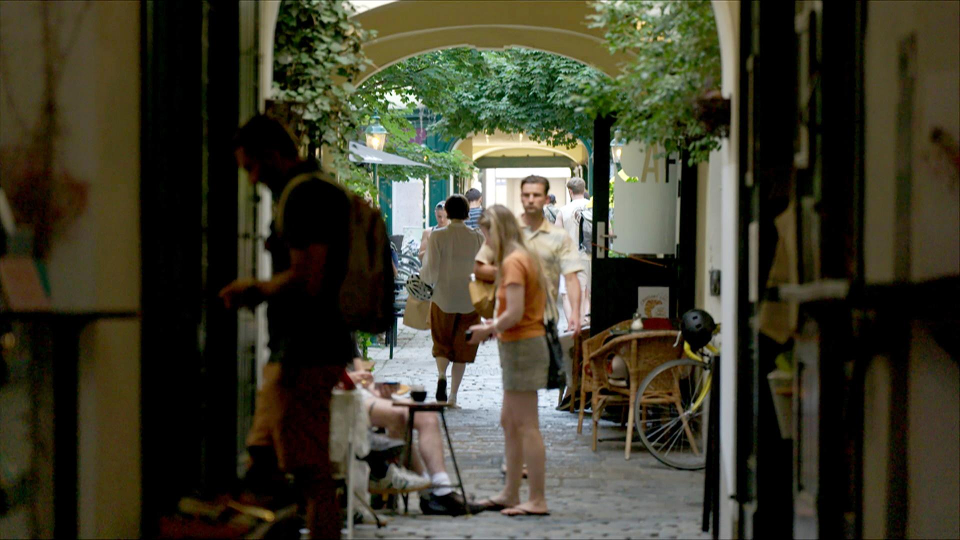 Das Bild zeigt einen belebten Innenhof in Wien, der von hohen Wänden umgeben ist. Über dem Kopf sind schmale, gewölbte Bögen zu sehen, die den Eingang zu einem Durchgang bilden. Im Vordergrund stehen mehrere Personen: Eine Frau mit langen, blonden Haaren trägt ein orangefarbenes Oberteil und eine graue Shorts. Sie hält die Hände über einem kleinen Tisch, auf dem eine Tasse steht. Neben ihr sitzt ein Mann, der einen leichten Kaffee zu trinken scheint. Im Hintergrund gehen mehrere Menschen, darunter eine Frau mit einer hellen Bluse und Hosen.   Der Weg im Innenhof ist aus Steinen gepflastert, und es gibt einige grüne Pflanzen, die das Bild lebendig wirken lassen. An den Wänden sind einige Lampen und dekorative Elemente sichtbar. Auf der linken Seite ist ein Teil eines Fahrrads mit einem gelben Helm zu sehen, der an einem Stuhl lehnt. Die Atmosphäre wirkt entspannt und einladend, abseits des städtischen Trubels.