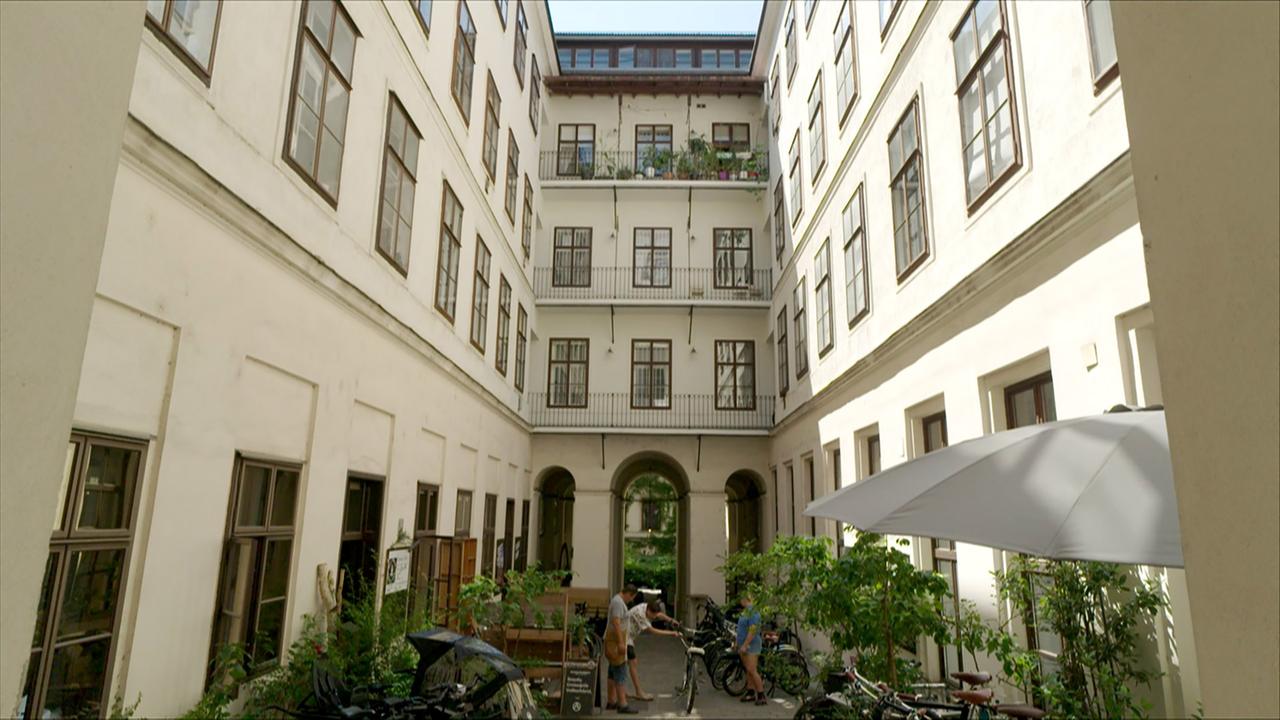 Das Bild zeigt einen Innenhof in Wien, genauer gesagt in einem Durchhaus im 7. Bezirk. Der Innenhof ist von mehrstöckigen Wohngebäuden umgeben, die helle, weiße Fassaden mit vielen Fenstern haben. Die Fenster verfügen über dunkelbraune Rahmen.   In der Mitte des Innenhofs stehen einige Fahrräder abgestellt. Auf der linken Seite gibt es eine Holzbank und mehrere Pflanzen, die den Hof begrünen. Außerdem ist ein Sonnenschirm aufgestellt, der Schatten spendet. Zu sehen sind auch zwei Personen, eine Frau und ein Mann, die sich um ein Fahrrad kümmern. Der Boden des Innenhofs ist gepflastert, und durch einen Bogen in der Mitte ist der Zugang zu einem weiteren Bereich sichtbar.   Das Licht scheint hell zu sein, was auf einen sonnigen Tag hinweist. Der Ort vermittelt eine ruhige, einladende Atmosphäre, die typisch für viele versteckte Innenhöfe in Wien ist.