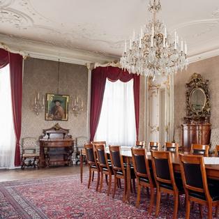 Das Bild zeigt einen eleganten, opulent gestalteten Raum im Augartenpalais. Der Raum ist groß und hat hohe Decken mit aufwendigen Verzierungen, die filigrane Stuckarbeiten aufweisen. An der Decke hängt ein großer, glitzernder Kronleuchter.  Die Wände sind mit einem zarten Muster in einem hellen Farbton dekoriert, und in einer der Wände ist ein großes, gerahmtes Porträt eines Mannes zu sehen. Flankiert von zwei Wandlampen ist dieser Bereich besonders hervorgehoben.   Vor dem Porträt befindet sich eine Holzkommode. Der Boden des Raumes ist mit einem persischen Teppich in warmen Rottönen ausgelegt. In der Mitte des Raumes steht ein großer, rechteckiger Tisch, umgeben von mehreren Holzstühlen mit schwarzen Sitzflächen.   Die Fenster sind mit schweren, roten Vorhängen dekoriert, die den Raum zusätzlich einladend wirken lassen. Auf der gegenüberliegenden Seite gibt es einen Kamin mit einem weiß verzierten Mantel und eine große, elegant geschwungene Holzvitrine, die antike Gegenstände beherbergt. Das gesamte Ambiente strahlt einen historischen und luxuriösen Charakter aus.