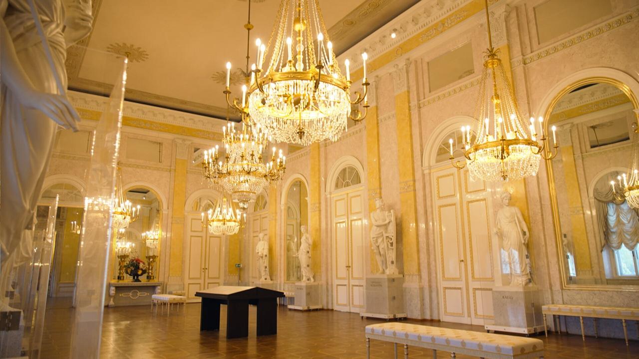 Das Bild zeigt den Innenraum des Albertina Saals in Wien. Der Raum ist prächtig gestaltet, mit hohen Decken und eleganten Wänden, die in sanften Gelbtönen gehalten sind. In der Mitte des Raumes hängen mehrere große Kristalllüster, die sanftes Licht verbreiten. An den Wänden stehen mehrere marmorne Statuen, die historische Personen darstellen. Der Boden besteht aus dunklem Holz und ist mit hellen Sitzbänken ausgestattet, die an den Wänden entlang stehen. Große Fenster mit eleganten Vorhängen sorgen für zusätzliches Licht und bieten Ausblick auf den Außenbereich. Die Atmosphäre des Raumes ist opulent und hebt die historische Bedeutung des Ortes hervor.