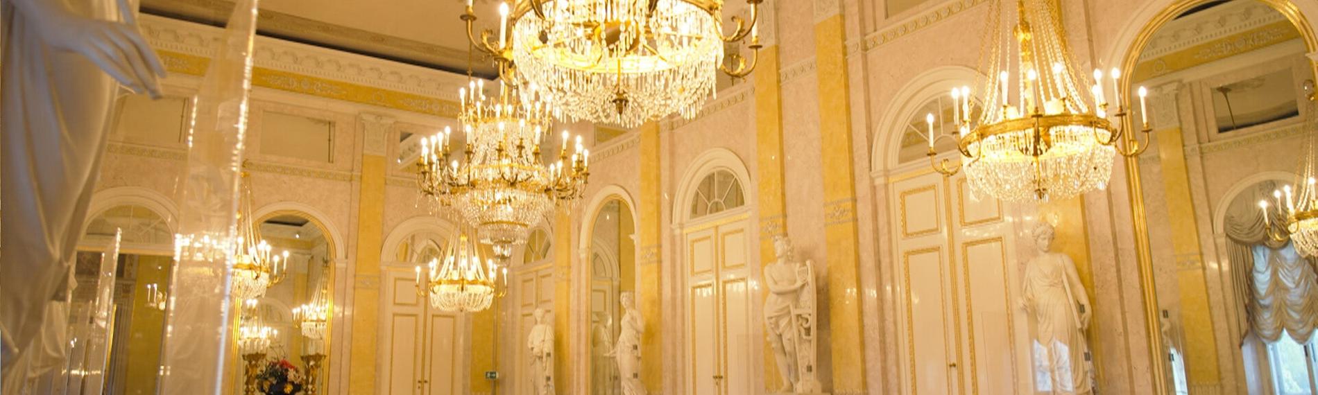 Das Bild zeigt den Innenraum des Albertina Saals in Wien. Der Raum ist prächtig gestaltet, mit hohen Decken und eleganten Wänden, die in sanften Gelbtönen gehalten sind. In der Mitte des Raumes hängen mehrere große Kristalllüster, die sanftes Licht verbreiten. An den Wänden stehen mehrere marmorne Statuen, die historische Personen darstellen. Der Boden besteht aus dunklem Holz und ist mit hellen Sitzbänken ausgestattet, die an den Wänden entlang stehen. Große Fenster mit eleganten Vorhängen sorgen für zusätzliches Licht und bieten Ausblick auf den Außenbereich. Die Atmosphäre des Raumes ist opulent und hebt die historische Bedeutung des Ortes hervor.