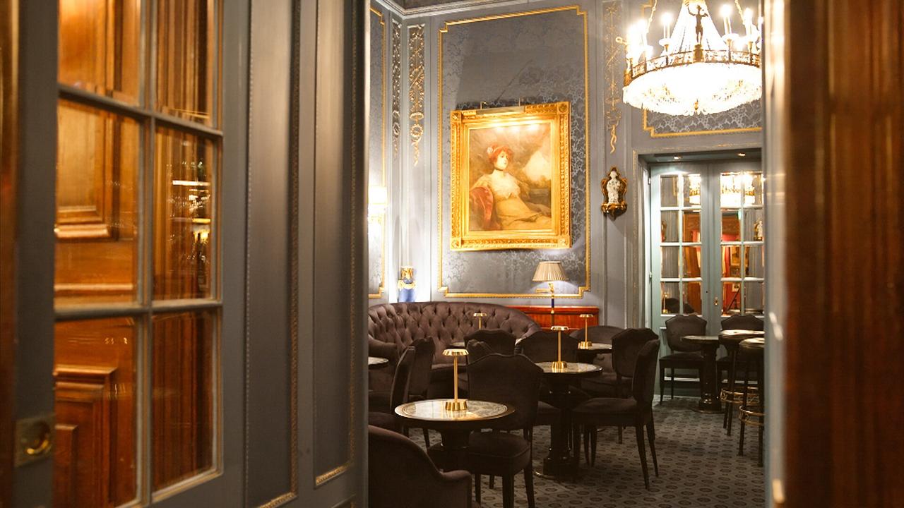 Das Bild zeigt einen eleganten Innenraum, der an das Ambiente eines stilvollen Cafés oder Salons erinnert. Der Raum hat eine geschmackvolle, historische Einrichtung mit einer Kombination aus blauen Wänden, goldenen Verzierungen und polierten Holzoberflächen.   Im Vordergrund sind mehrere kleine, runde Tische mit schwarzen Stühlen umgeben. Auf jedem Tisch steht eine kleine, goldfarbene Lampe. An der Wand hängt ein großes Gemälde mit einem majestätischen Portrait einer Frau, eingerahmt in einen prunkvollen goldenen Rahmen.   Eine luxuriöse Kristallchandelier beleuchtet den Raum, der somit eine gediegene Atmosphäre ausstrahlt. Im Hintergrund sind zwei große, verglaste Türen, die zu einem weiteren Raum führen. Der Teppichboden hat ein leicht gemustertes Design, das harmonisch zur restlichen Einrichtung passt.   Der Blick vermittelt den Eindruck von Intimität und Eleganz, ideal für gesellschaftliche Zusammenkünfte oder private Treffen.