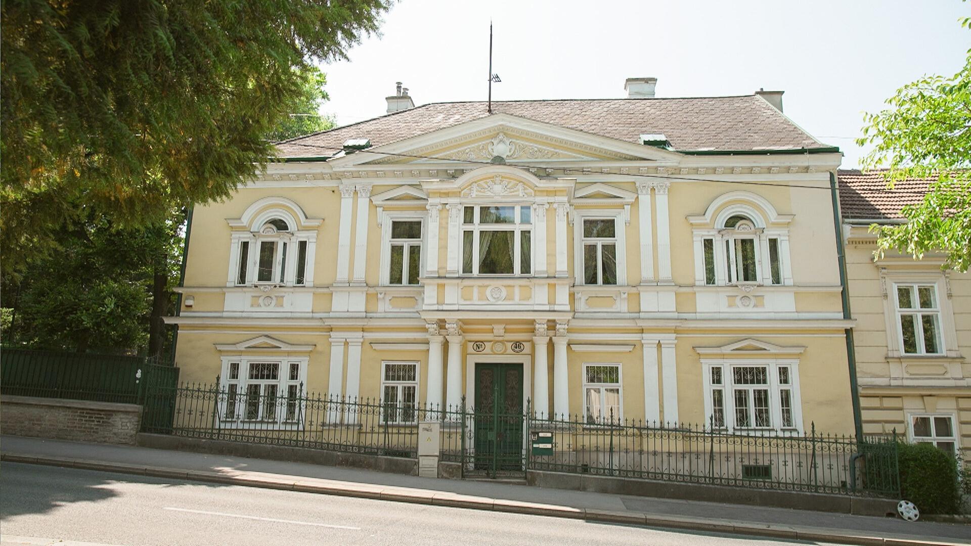 Das Bild zeigt die Fassade einer eleganten Villa in Hietzing, einem Stadtteil von Wien. Die Villa hat eine gelbe Außenwand mit weißen, kunstvollen Verzierungen, die den klassischen Stil des 19. Jahrhunderts repräsentieren.   In der Mitte der Fassade befindet sich der Eingang, der durch eine grüne Tür markiert ist. Über der Tür sind zwei große Fenster, die symmetrisch angeordnet sind. Weitere Fenster sind entlang der Fassade verteilt, einige mit Rundbögen.   Die Villa ist von einem niedrigen, grünen Zaun umgeben, der sich entlang der Straße erstreckt. Vor der Villa stehen einige hohe Bäume, die einen Teil der Sicht auf das Gebäude verdecken. Der Himmel ist klar und der Bereich insgesamt wirkt ruhig und wohlhabend.   In der EXIF-Beschreibung wird erwähnt, dass die Villa eine bedeutende historische Rolle spielt, da hier Kaiser Franz Joseph seine Geliebte besuchte.