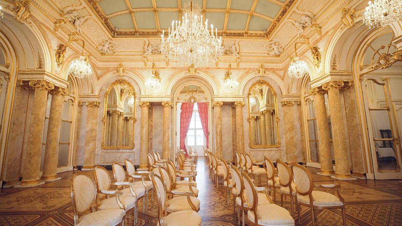 Das Bild zeigt einen opulent dekorierten Raum im Palais Coburg in Wien. Die Wände sind mit hellem Marmor verkleidet, und goldene Verzierungen ziehen sich durch die gesamten Mauern und Decken. Die Decke ist kunstvoll gestaltet und hat eine quadratische Struktur mit zarten reliefartigen Elementen.  In der Mitte des Raumes hängen mehrere Kristalllüster, die für eine glamouröse Beleuchtung sorgen. An den Seiten des Raumes sind hohe Fenster mit roten Vorhängen, die den Raum zusätzlich aufhellen. Der Boden ist mit einem kunstvollen Holzparkett verziert, das komplizierte Muster aufweist.  Entlang der Wände stehen elegant gestaltete Stühle mit polierten Holzrahmen und hellen, gepolsterten Sitzflächen. Der Raum vermittelt eine Atmosphäre von Luxus und historischer Bedeutung, die die aristokratische Vergangenheit des Palais widerspiegelt.