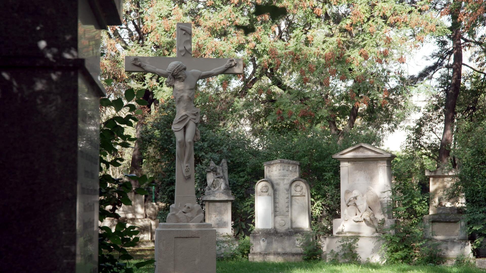 Das Bild zeigt einen Friedhof im Waldmüller Park in Wien. Im Vordergrund steht ein weißer Kruzifix-Skulptur, die Jesus am Kreuz darstellt. Die Figur ist auf einem Sockel platziert und wird von üppigem Grün umgeben. Im Hintergrund sind mehrere Grabsteine zu sehen, die unterschiedliche Formen und Designs aufweisen. Einige Grabsteine sind schlicht gehalten, während andere detaillierte Reliefs oder Skulpturen zeigen, wie zum Beispiel eine Figur eines weinenden Engels. Die Umgebung ist von hohen Bäumen und strauchartiger Vegetation geprägt, die teilweise das Licht filtern und eine ruhige, melancholische Atmosphäre schaffen. Die Gesamtstimmung des Bildes vermittelt ein Gefühl der Erinnerung und der Ruhe.