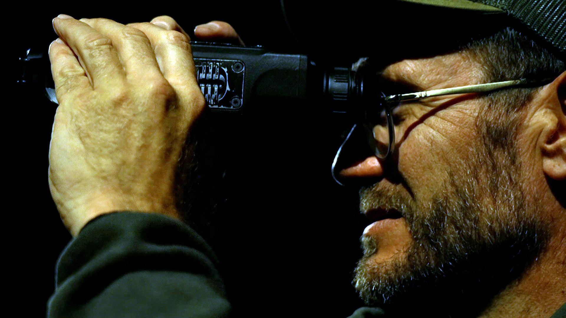 Nahaufnahme eines Mannes im Profil, der nachts durch ein Fernglas schaut. Er trägt eine Brille und eine Mütze, und seine Hand hält das Fernglas fest, während er konzentriert in die Dunkelheit blickt.