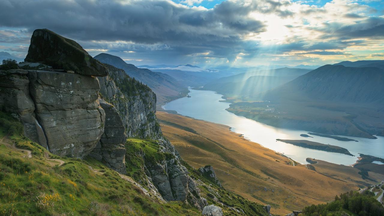 Eine Landschaft der schottischen Highlands. Im Vordergrund einige Felsen in Graslandschaft und im Hintergrund Berge und Gewässer.