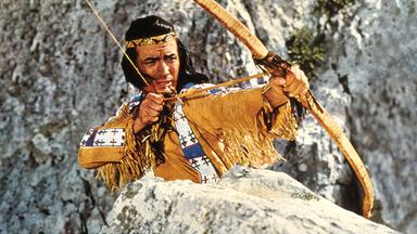 Großaufnahme von Winnetou (Pierre Brice) mit einem gespannten Bogen vor einer Felslandschaft.