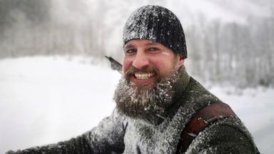 Nahaufnahme: Ein Mann mit Vollbart und Mütze steht lachend im Schnee. 