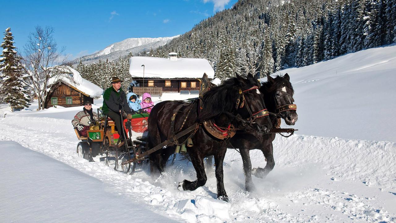 Vor blauem Himmel an dick verschneiten Berghütten vorbei in einem bunten Schlitten, der von zwei Pferden gezogen wird