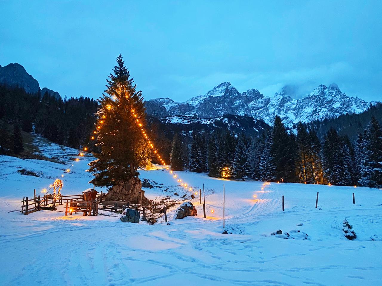 Das Bild zeigt eine Winterlandschaft in Filzmoos, einer malerischen Region im Pongau. Im Vordergrund steht ein großer, beleuchteter Weihnachtsbaum, um den Lichterketten in Form eines großen Dreiecks angebracht sind. Der Baum ist umgeben von einem verschneiten Gelände, das sanft angehobene Hügel und Tannenbäume umfasst. Die Schneedecke ist gleichmäßig und frisch, was der Szene ein idyllisches Aussehen verleiht.   Im Hintergrund erheben sich beeindruckende Berggipfel, die teilweise von Wolken umhüllt sind. Die Stimmung ist ruhig und beschaulich, und der Himmel zeigt eine sanfte Dämmerung mit bläulichen und grauen Tönen. Entlang des Weges sind kleine Lichtquellen sichtbar, die Wärme und Gemütlichkeit in die winterliche Umgebung bringen.   Das Bild vermittelt das Gefühl von Tradition und Naturverbundenheit in der Winterzeit.
