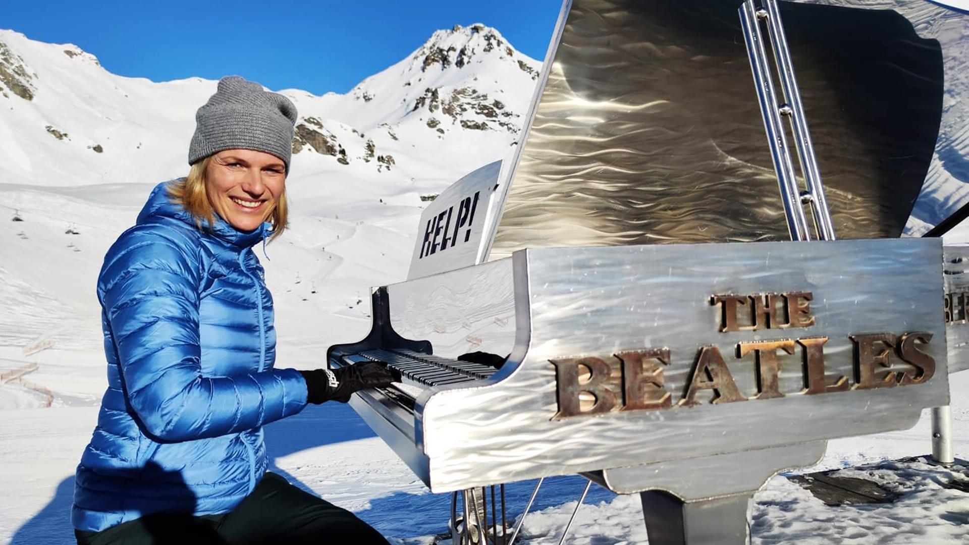 Eine winterlich gekleidete Frau spielt in einer verschneiten Berglandschaft an einem silbernen Flügel