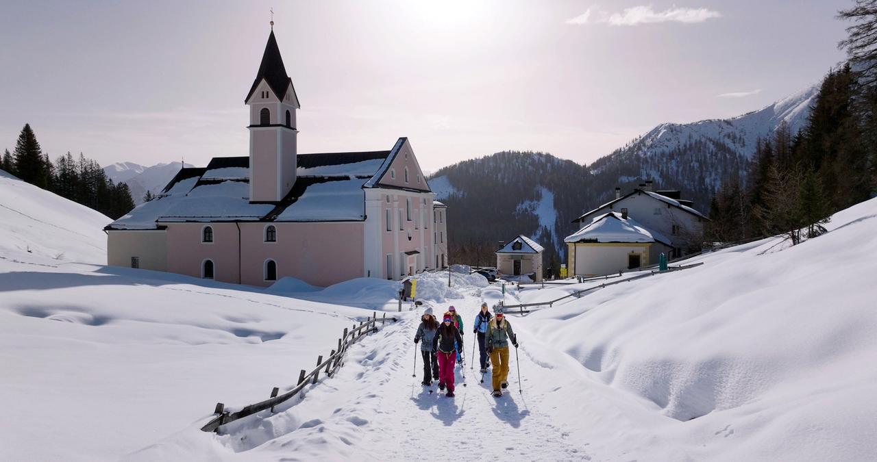 Das Bild zeigt eine winterliche Landschaft in den Tiroler Alpen, vermutlich im Wipptal. Im Zentrum steht ein pinkfarbenes Klostergebäude mit einem charakteristischen, spitzbübigen Turm, das von einer schneebedeckten Umgebung umgeben ist. Um das Kloster herum liegt eine sanft geschwungene Schneelandschaft mit Hügeln, die das Licht der Sonne reflektieren.   Eine Wandergruppe aus fünf Personen bewegt sich auf einem schmalen, verschneiten Weg in Richtung des Klosters. Sie tragen Winterkleidung und -ausrüstung, einige haben Skistöcke dabei. Ein Holzgeländer verläuft parallel zum Weg, und im Hintergrund sind schneebedeckte Berge sichtbar, die die Kulisse bilden. Der Himmel ist klar mit einem sanften Licht, das über die Szene strahlt.   Insgesamt vermittelt das Bild einen Eindruck von Ruhe und winterlicher Idylle in den Alpen.