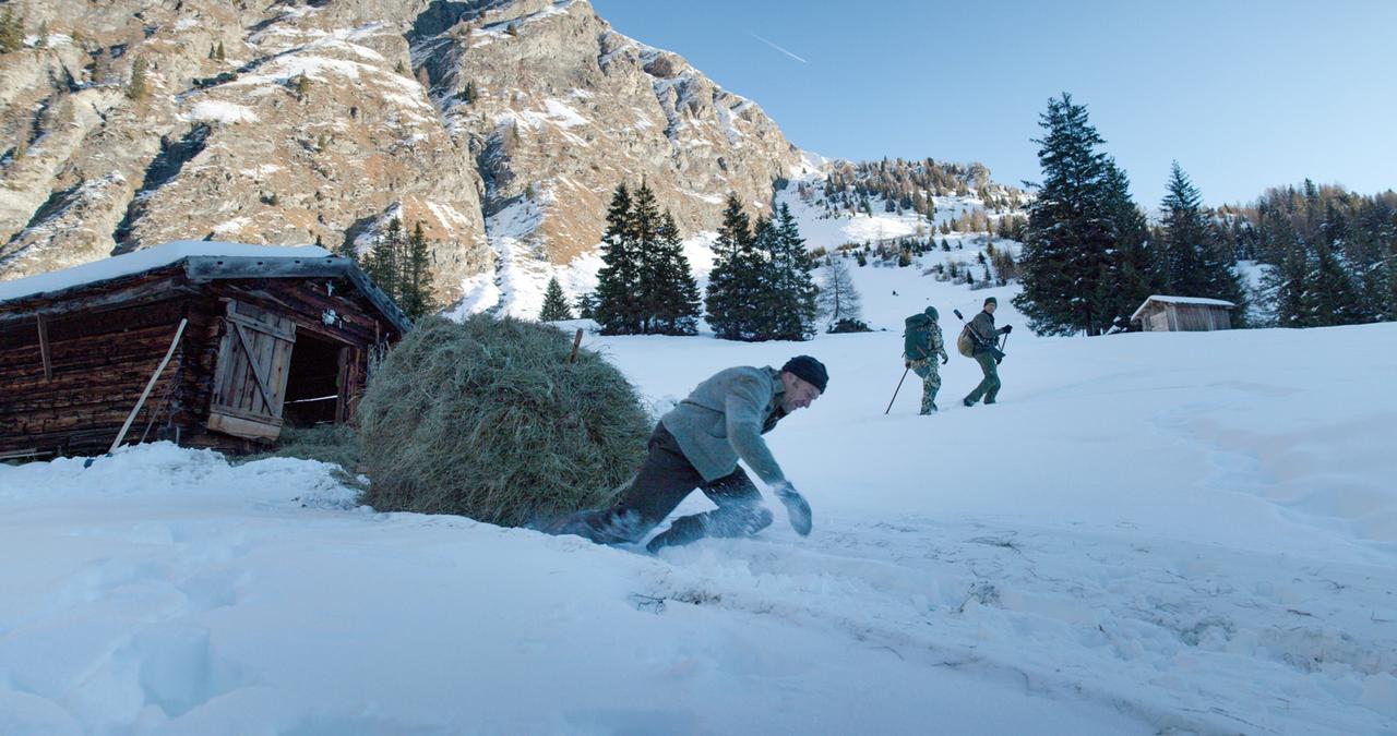 Das Bild zeigt eine winterliche Landschaft im Gebirge, vermutlich im Wipptal, wo eine Schneedecke den Boden bedeckt. Im Vordergrund ist ein Mann zu sehen, der im Schnee auf allen Vieren rutscht, während er eine große Heuballe hinter sich zieht. Er trägt einen grauen Wollpulli und eine schwarze Mütze. Auf der linken Seite des Bildes steht eine rustikale Holzhütte, die teilweise mit Schnee bedeckt ist.   Im Hintergrund erheben sich steile Felswände, die mit schneebedeckten Nadelbäumen durchzogen sind. Zwei weitere Männer sind rechts im Bild zu sehen; einer trägt einen grünen Rucksack und eine lange Waffe, während der andere einen weiteren Rucksack hat. Der Himmel ist klar und blau, was auf sonniges Wetter hinweist. Insgesamt vermittelt die Szene eine ruhige, abgelegene Atmosphäre in einer winterlichen Berglandschaft.