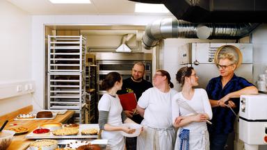 Zwei Männer und drei Frauen stehen in einer modernen Backstube. Sie blicken einander an und lächeln. Die drei Frauen haben weiße Konditoren-Arbeitskleidung an, eine hält eine Schüssel in der Hand, eine andere einen großen Schneebesen. An der Seite steht ein Tablett mit drei Kuchen.